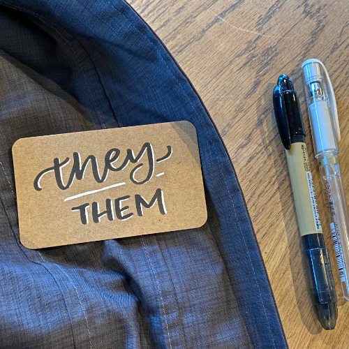 A kraft cardstock magnet pin lettered with they/them in brush pen